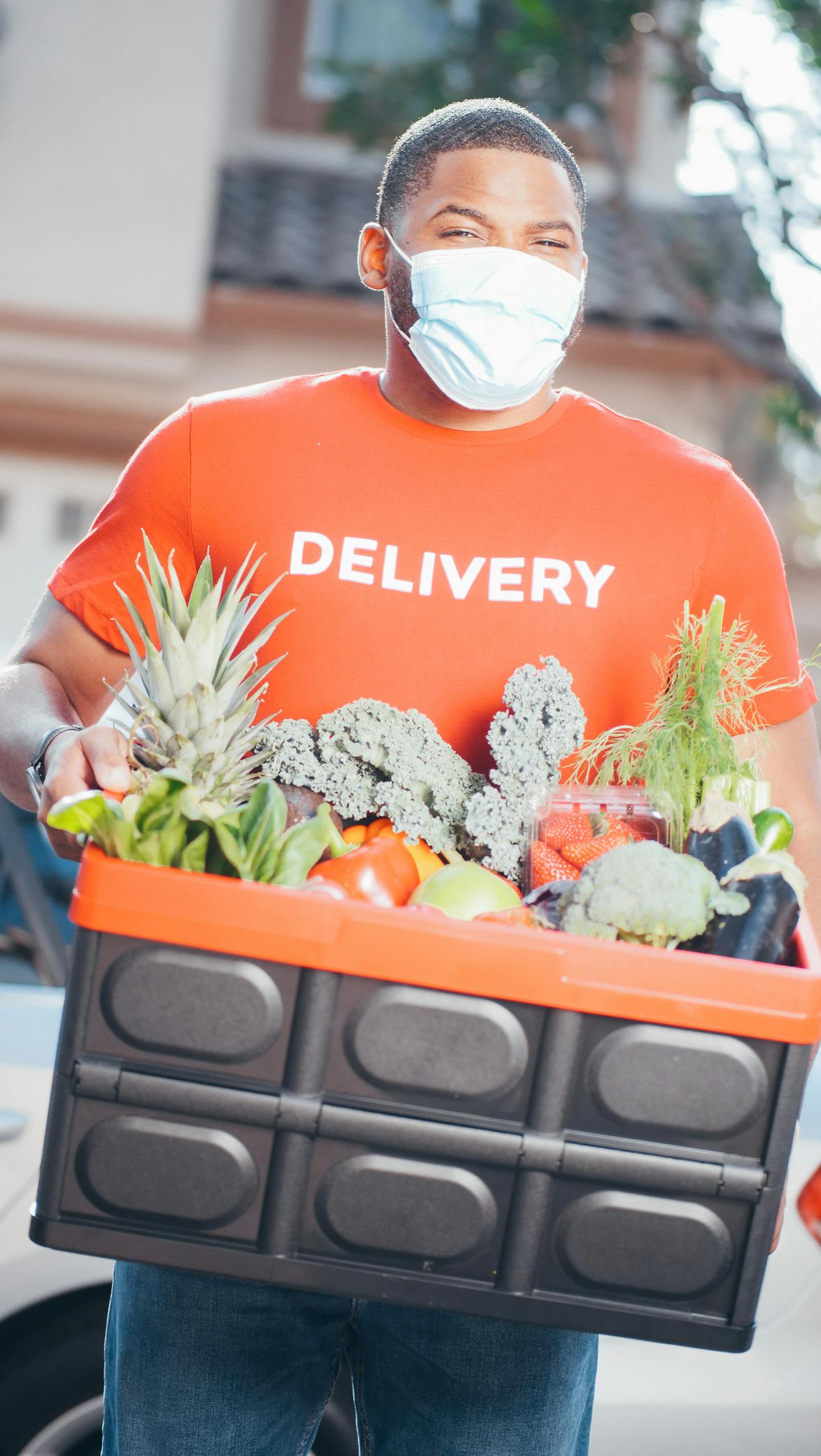 A delivery man wearing a face mask carries a box of fresh produce. Outdoor scene, health-focused.