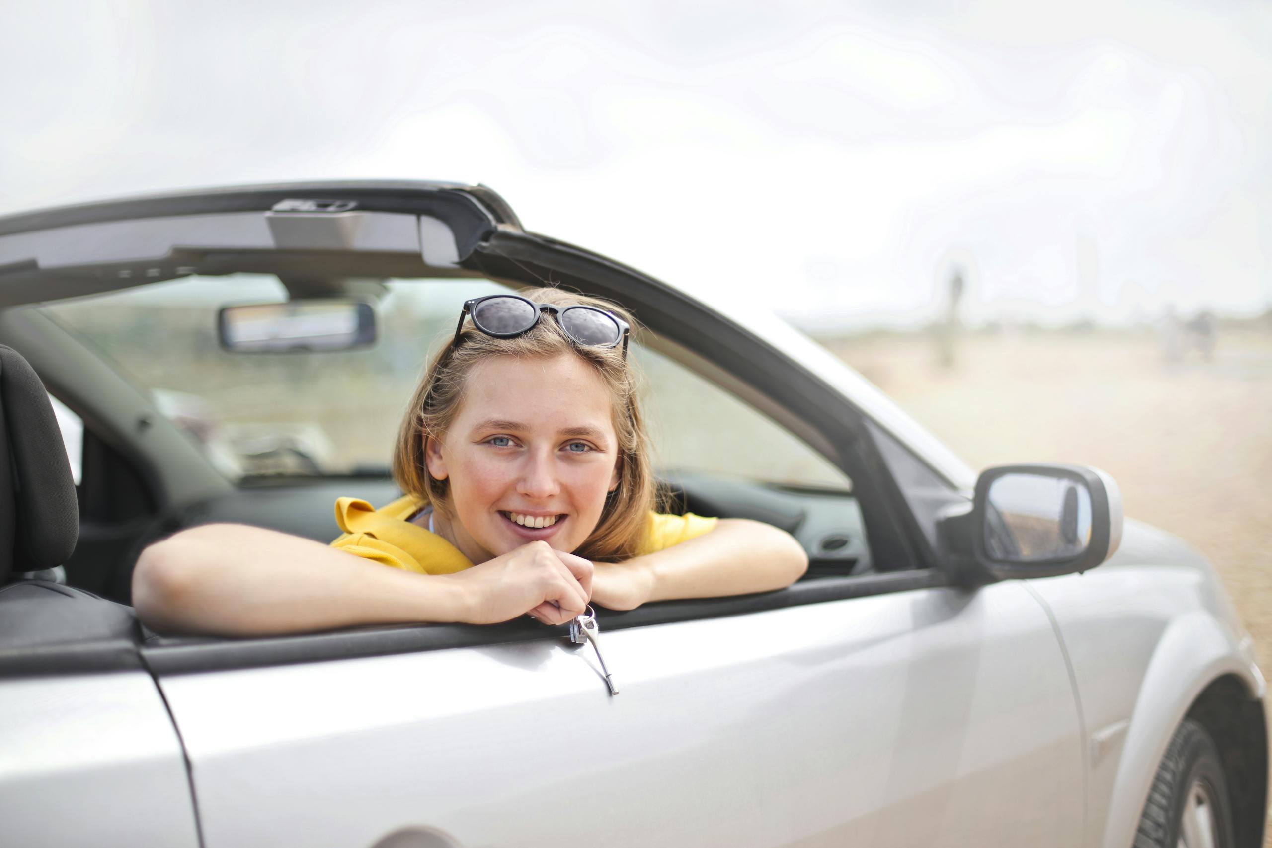 A young woman smiles joyfully while leaning out of a convertible car, enjoying a sunny day.