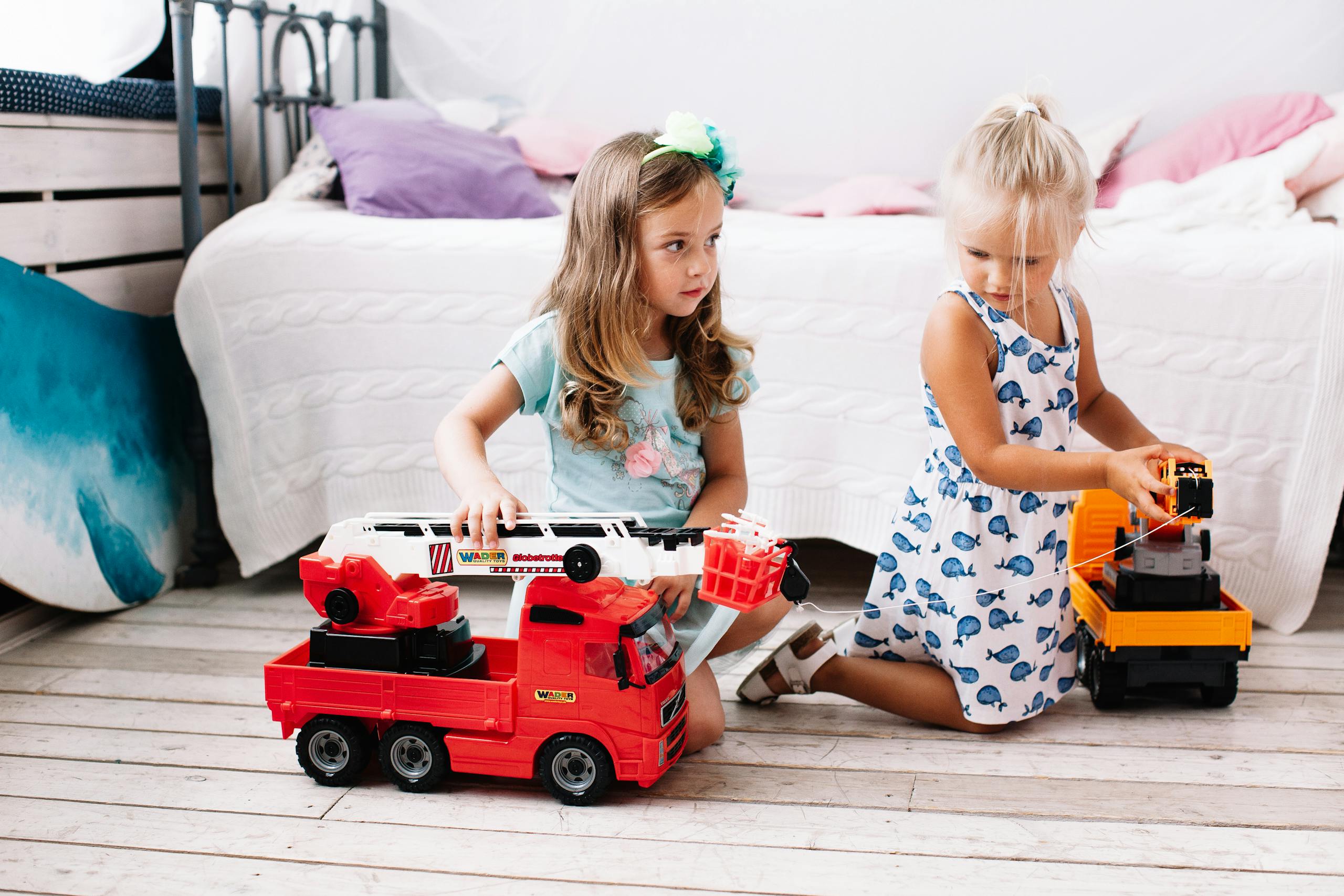 Two young girls playing with colorful toy trucks on a wooden floor in a cozy room.