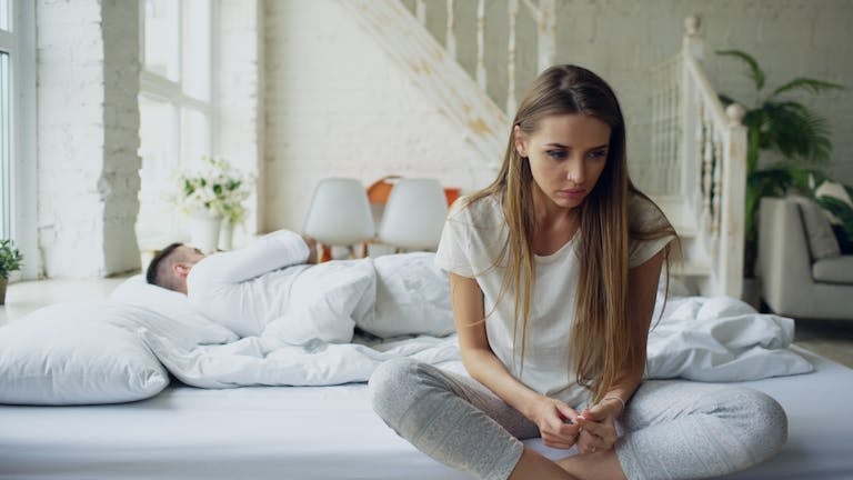 Woman sitting on bed looking thoughtful while man sleeps, symbolizing relationship tension.