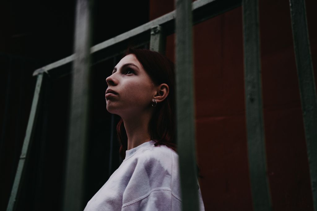 A woman in a white shirt with earrings looking up behind metal bars, creating a moody atmosphere.