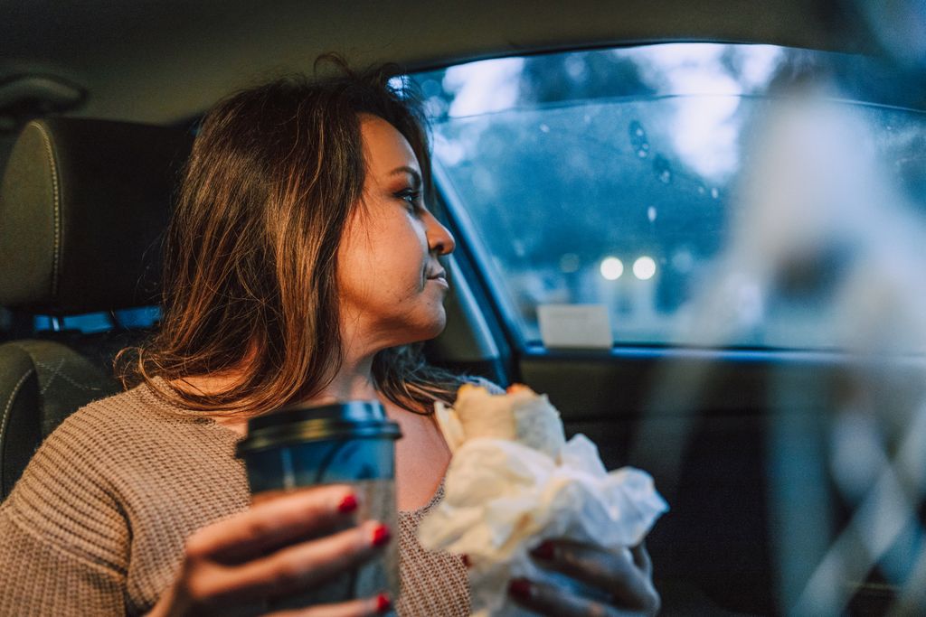 A woman sits in her car with a coffee and a sandwich, reflecting on her day.