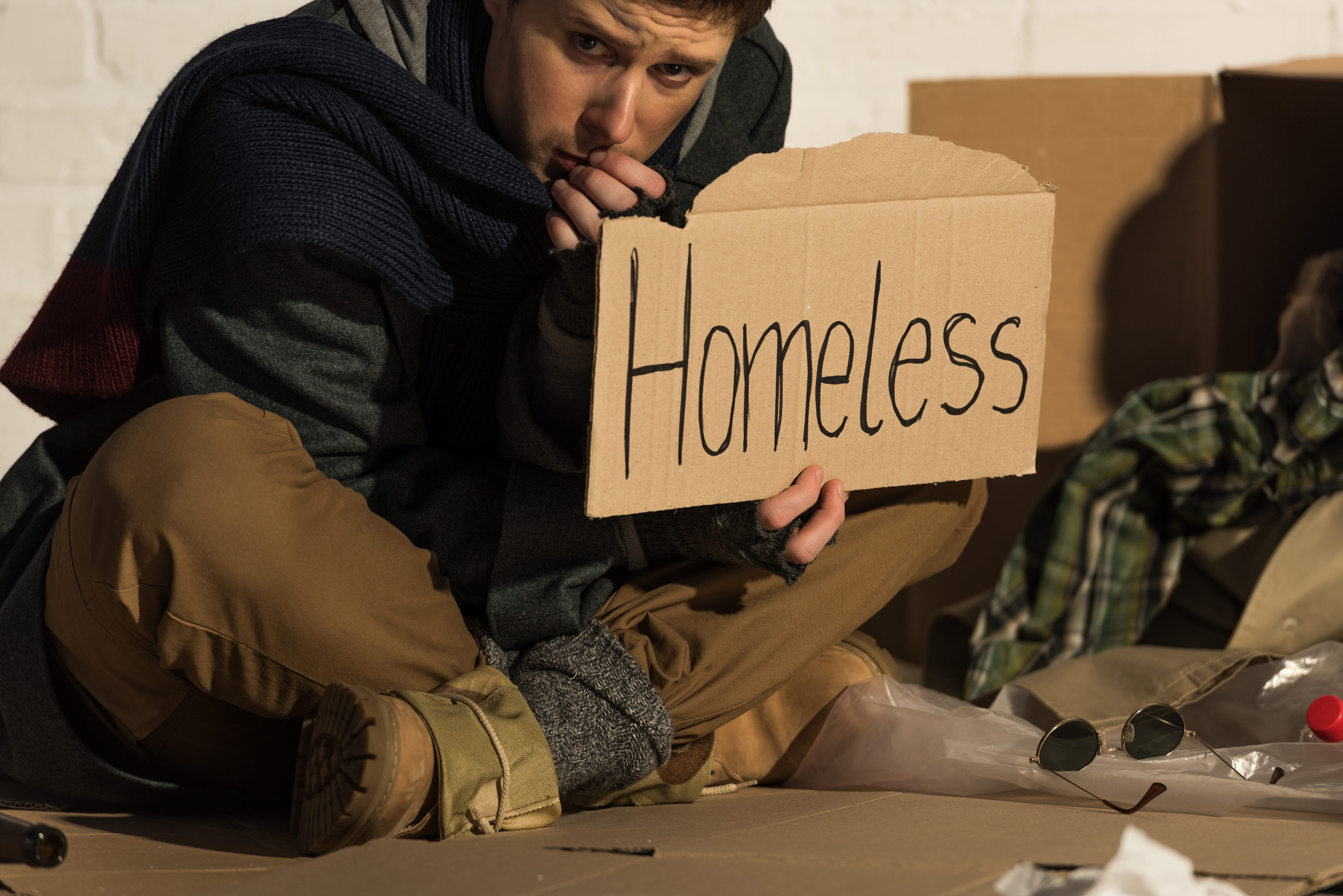 depressed man sitting on cardboard and holding card with "homeless" handwritten text