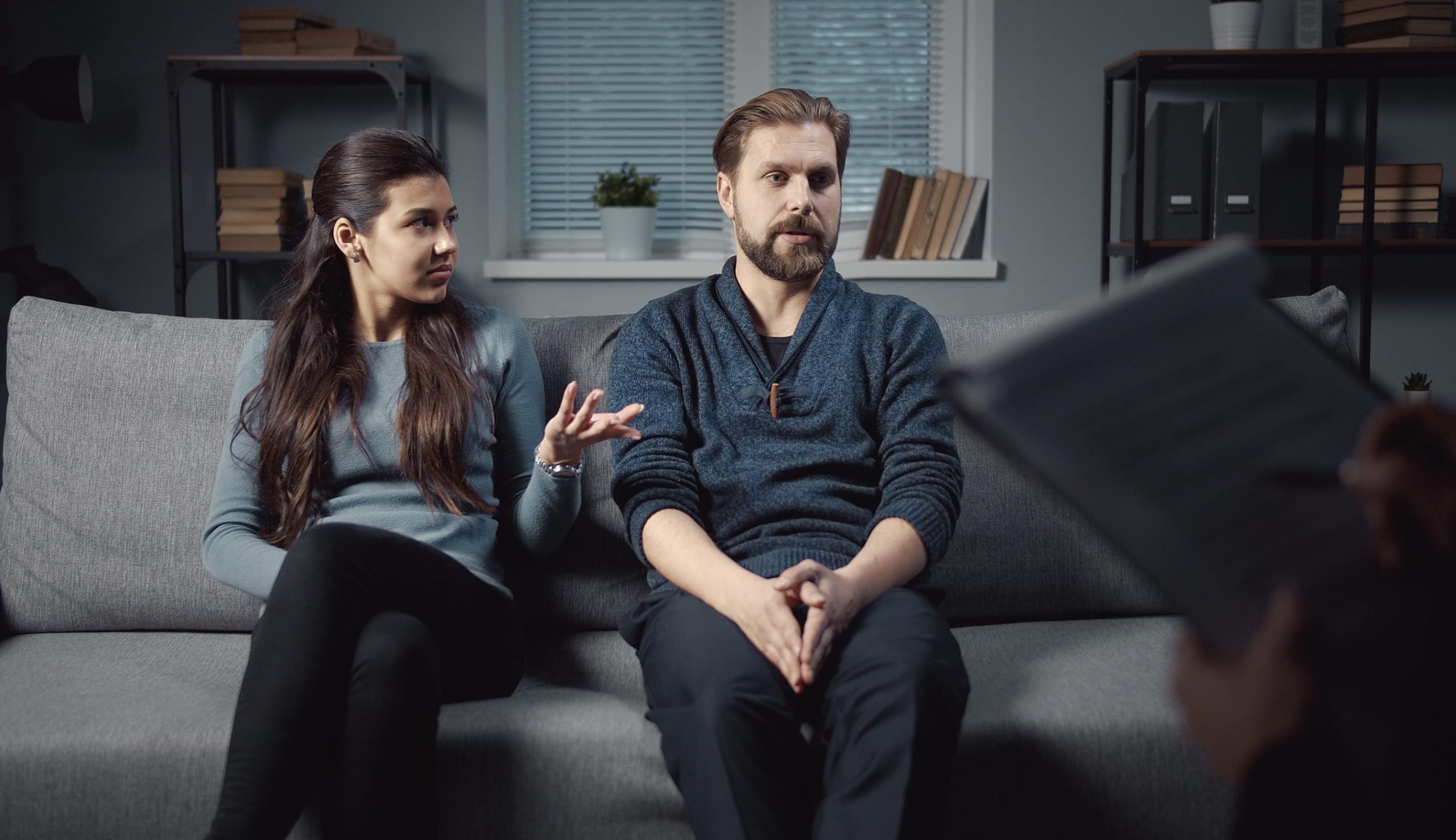 Couple sitting on sofa visiting psychologist to solve family problem, woman gestures, man speaks