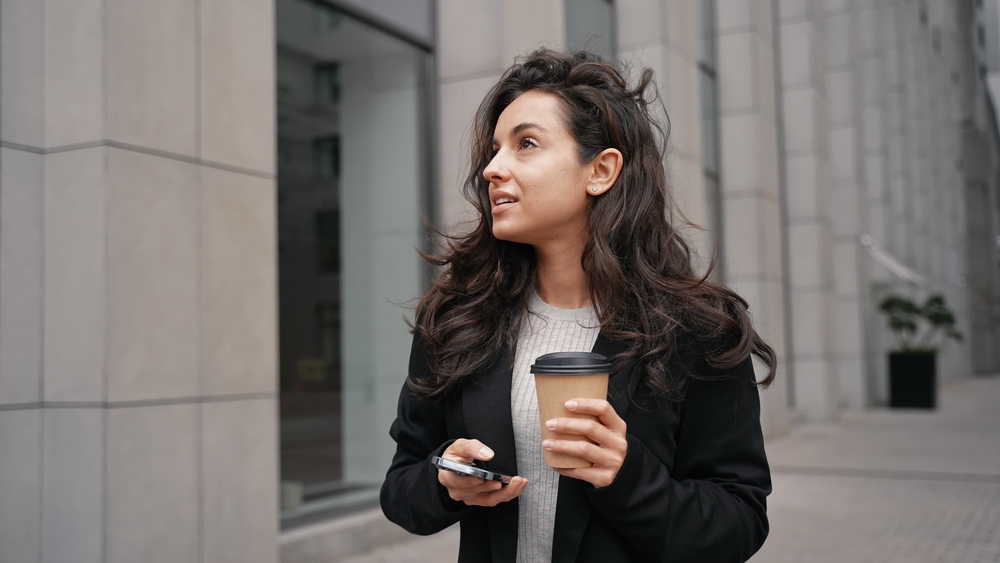 Young Caucasian woman walking in big city, looking around, admiring contemporary buildings in the city downtown. Female with dark long hair walking, smiling, looking at her phone, and drinking coffee.