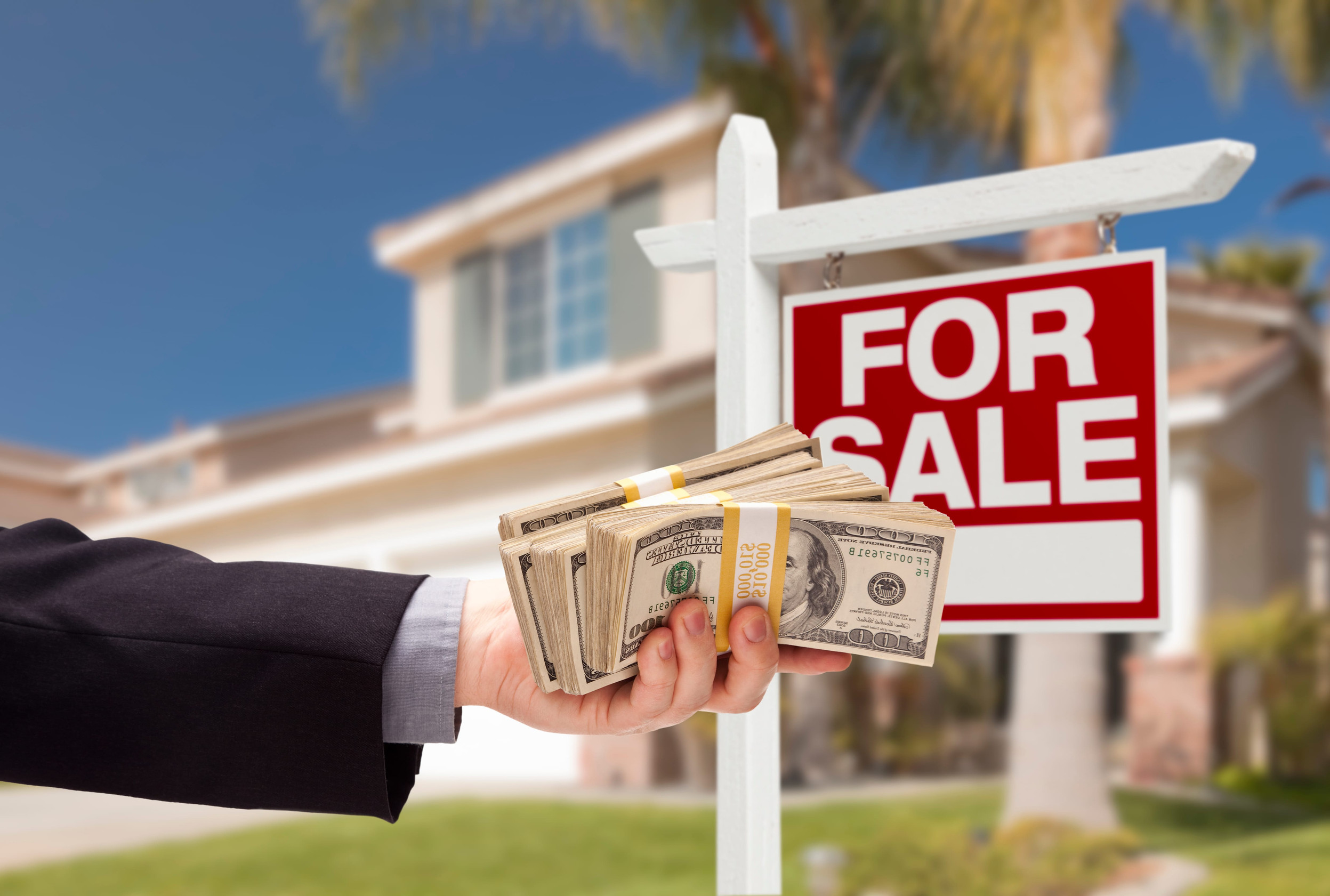 Buyer Handing Over Cash for House with Home and For Sale Real Estate Sign Behind.