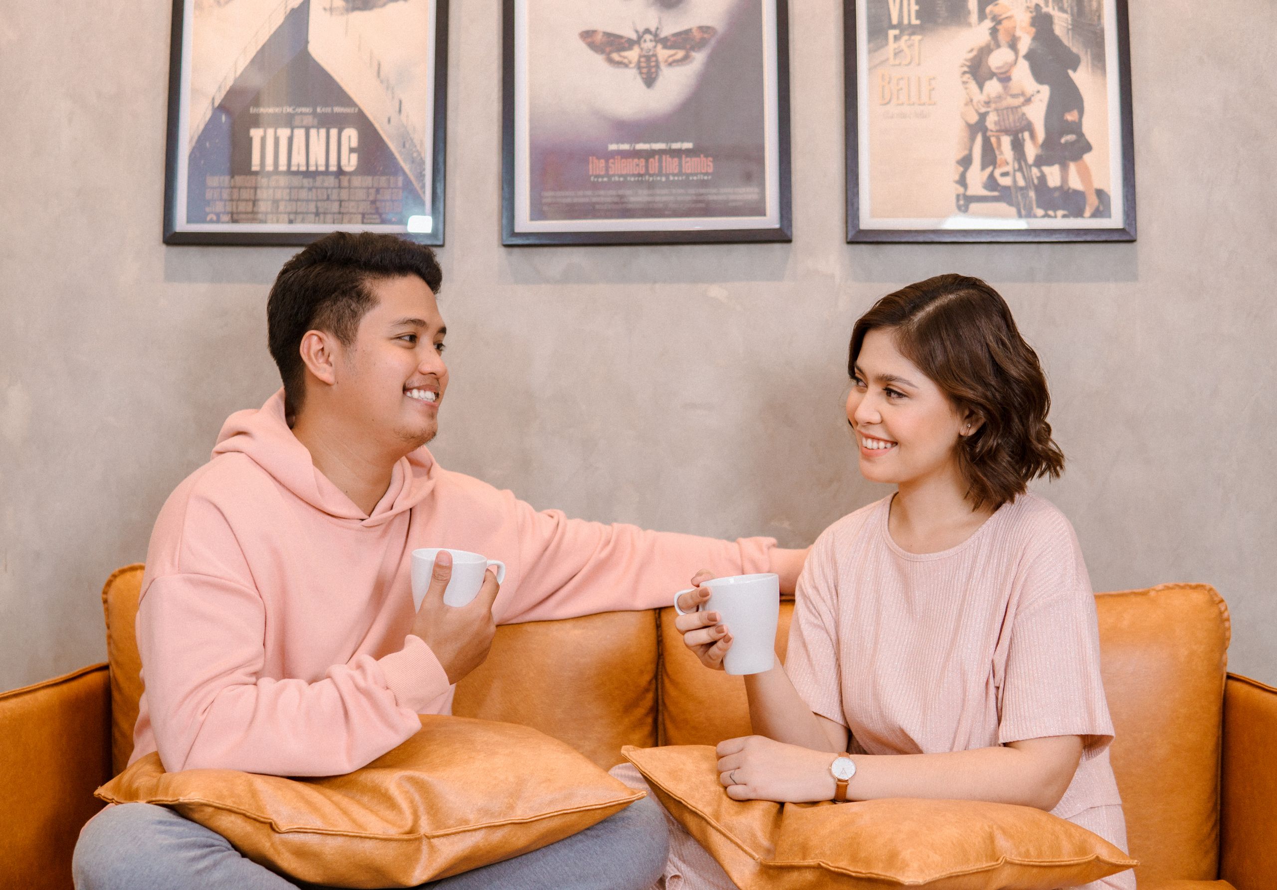 A cheerful couple in pink attire enjoys coffee on a cozy couch at home.