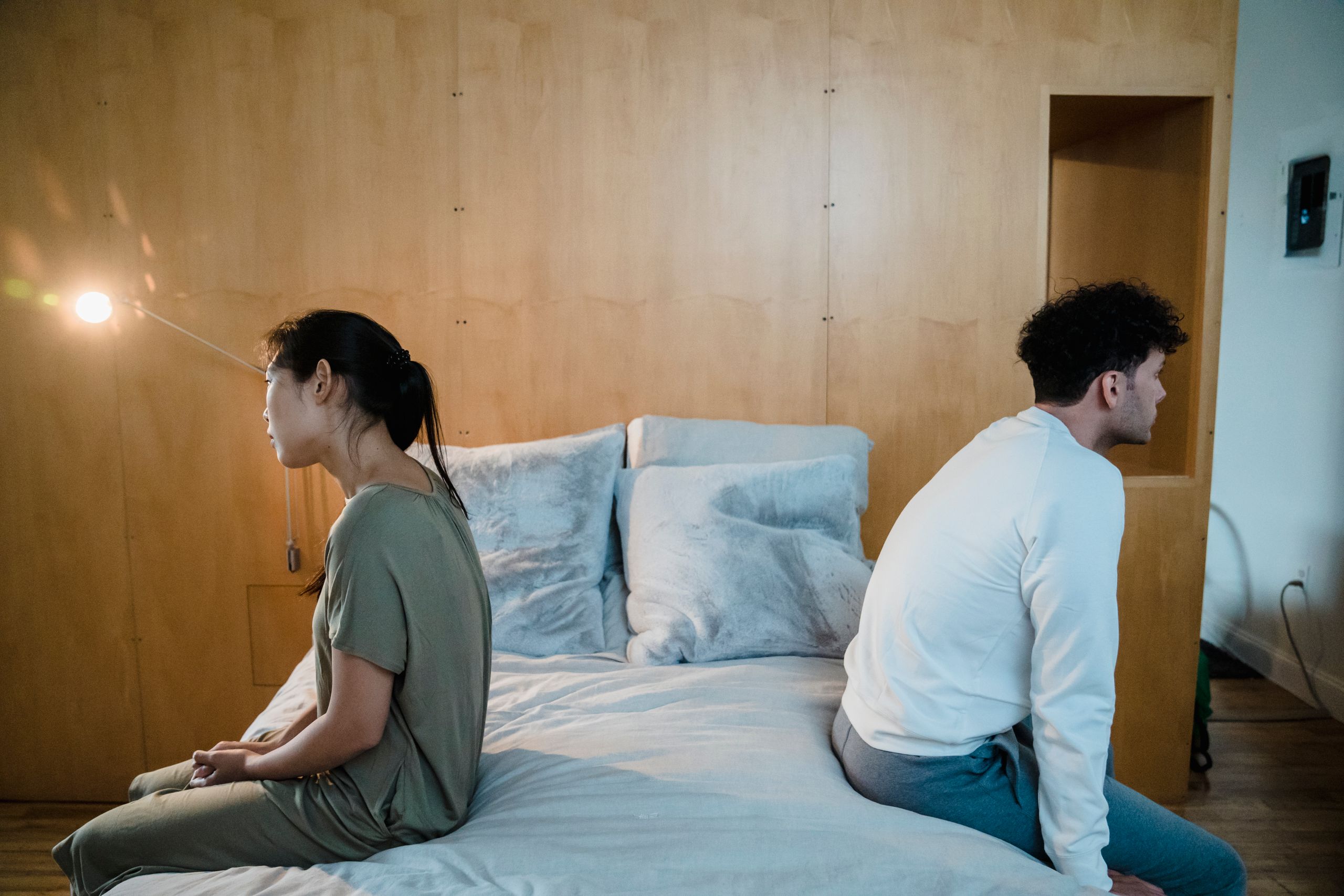 A couple sits back to back on a bed, showing tension and misunderstanding in a cozy bedroom.