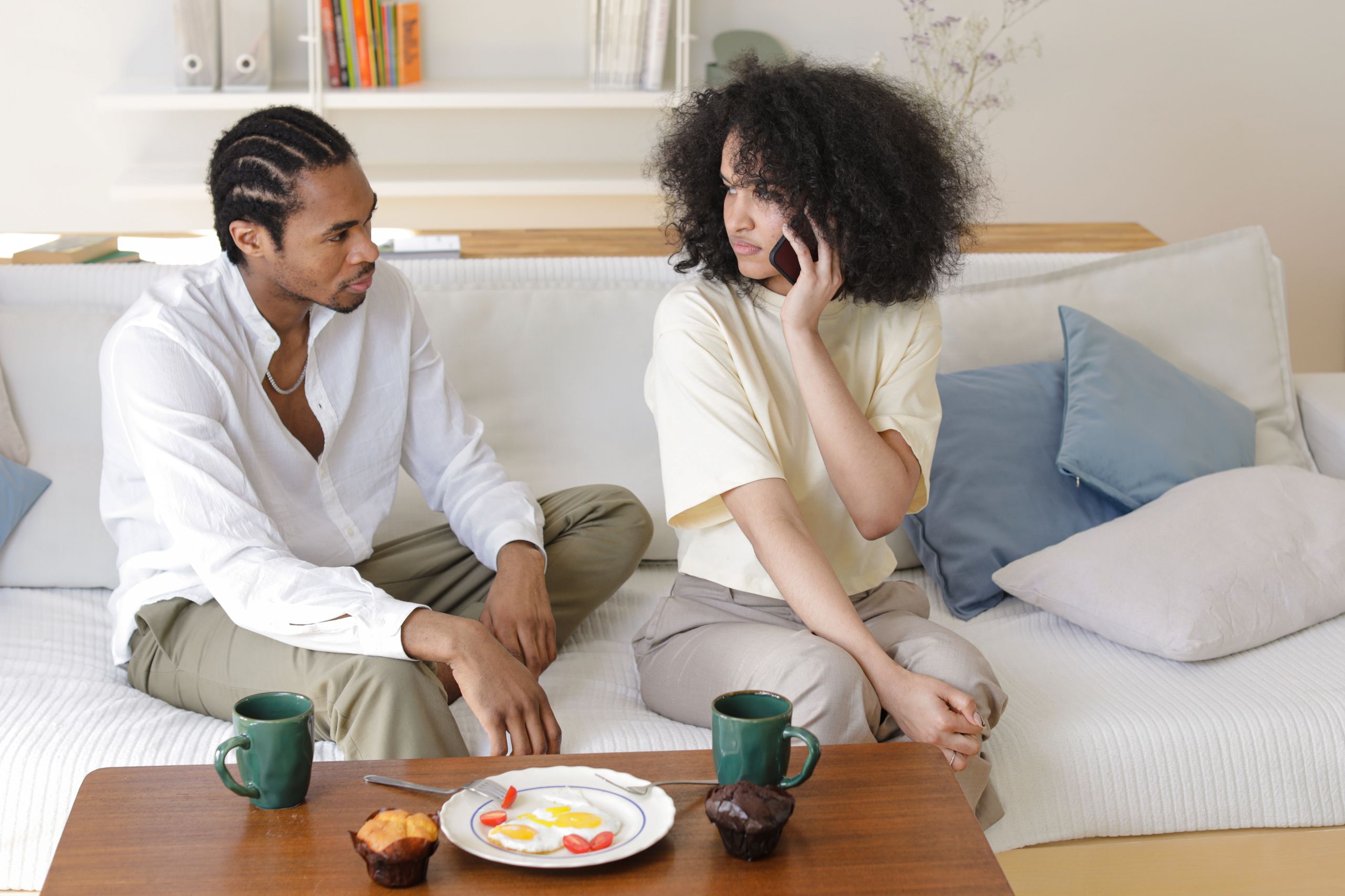 A couple sits on a sofa, having a conversation while one holds a smartphone indoors.