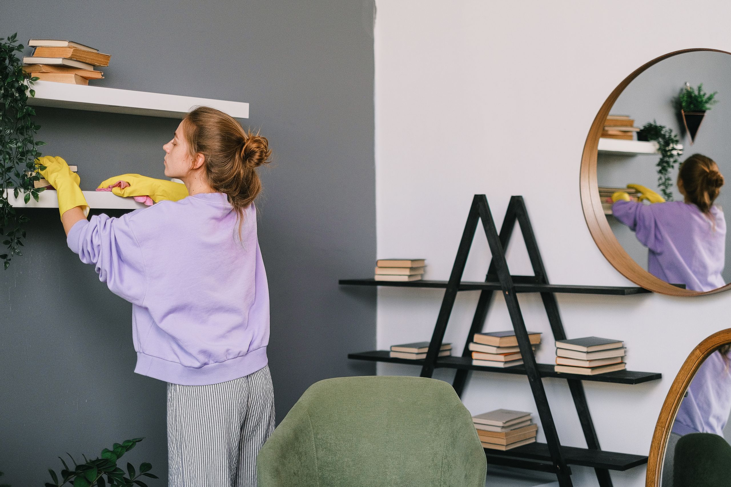 A woman cleans shelves in a stylish living room with mirrors and decor.