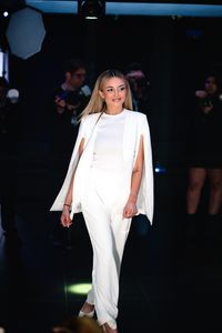 Blonde woman in elegant white suit smiles while walking on a fashion show runway.