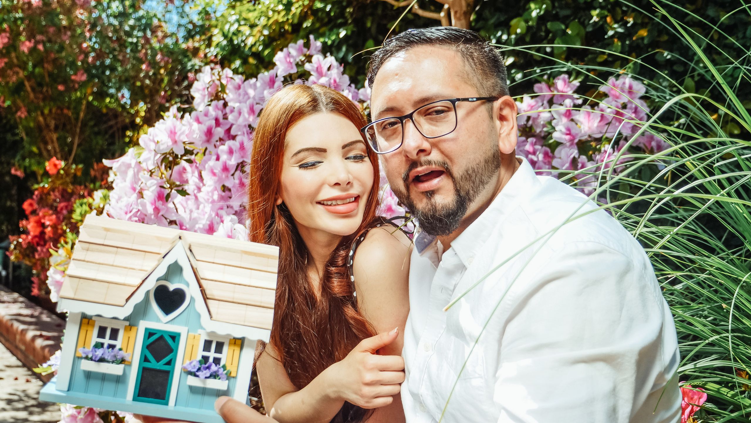 Cheerful couple holding a miniature house, symbolizing a new home purchase in a garden setting.