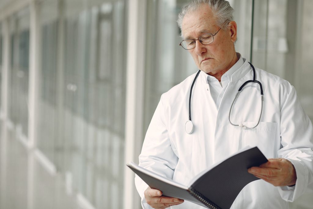 Crop concentrated professional senior male physician in white robe with stethoscope standing in modern hospital hallway and reading medical case records in daylight