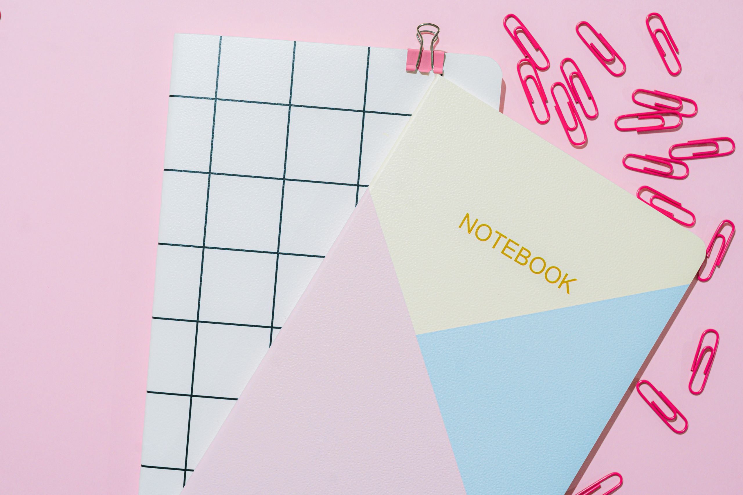 Flat lay of notebooks, paper clips, and binder clip on a pink background.