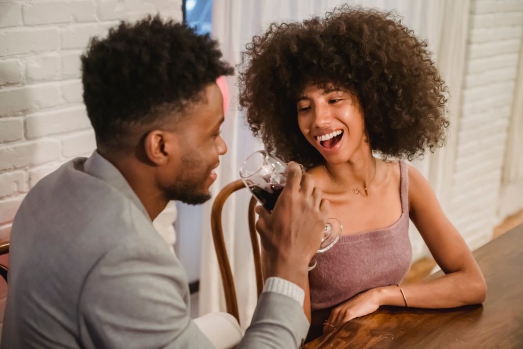 Happy young African American couple in elegant clothes drinking red wine from glasses while sitting at table in bright apartment near curtains and brick wall