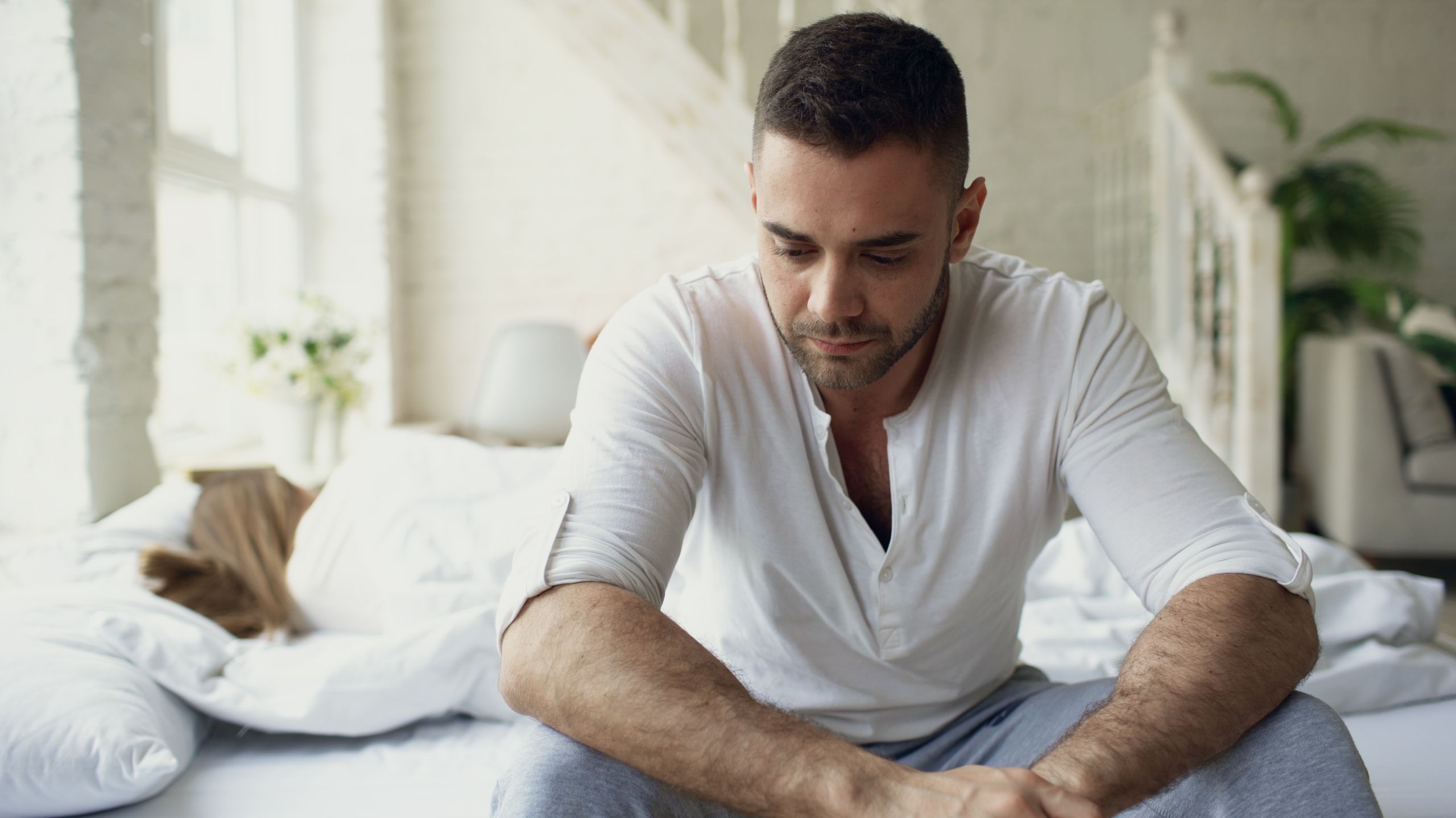 Man deep in thought while sitting on a bed in a sunlit bedroom.