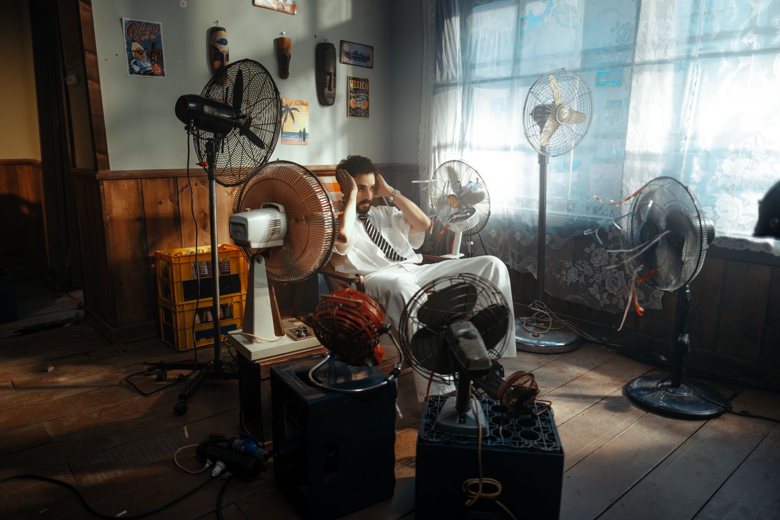 Man in white outfit sitting surrounded by several fans indoors during daytime.