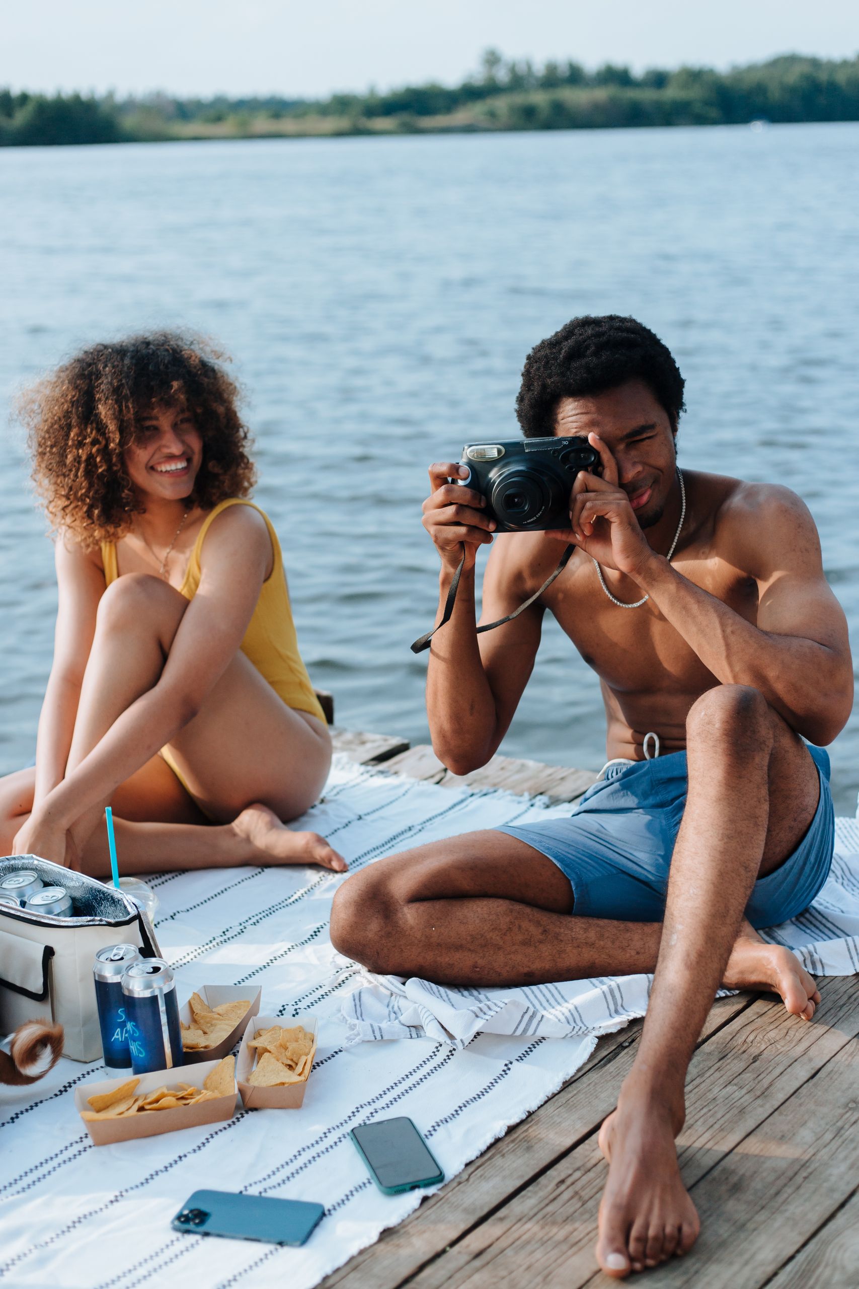 Two friends enjoying a summer picnic by the lake, one taking photos, capturing joyful moments.