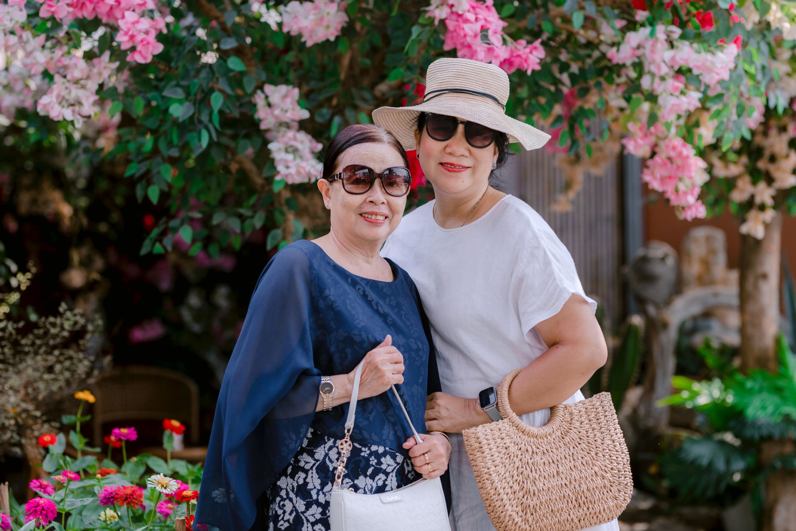 Two senior women smiling outdoors in a vibrant garden setting with pink flowers.