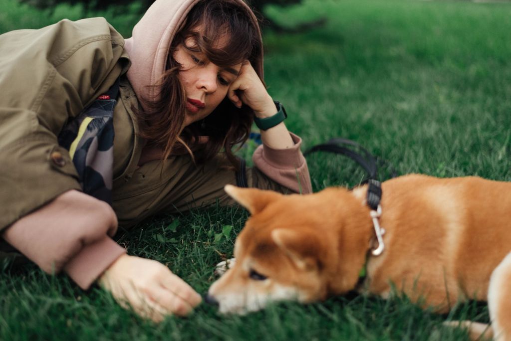 Woman in a jacket relaxing on grass with her Shiba Inu dog on a leash.