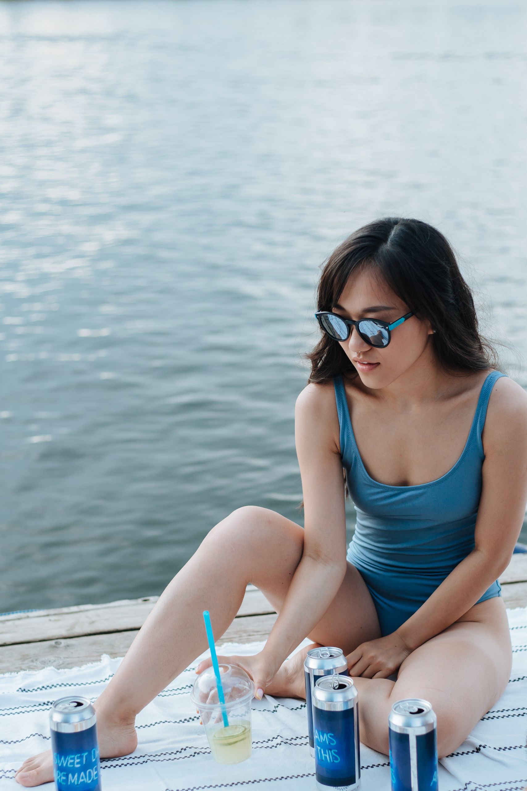 Woman relaxing at a lakeside picnic with drinks in summer, wearing sunglasses and swimwear.