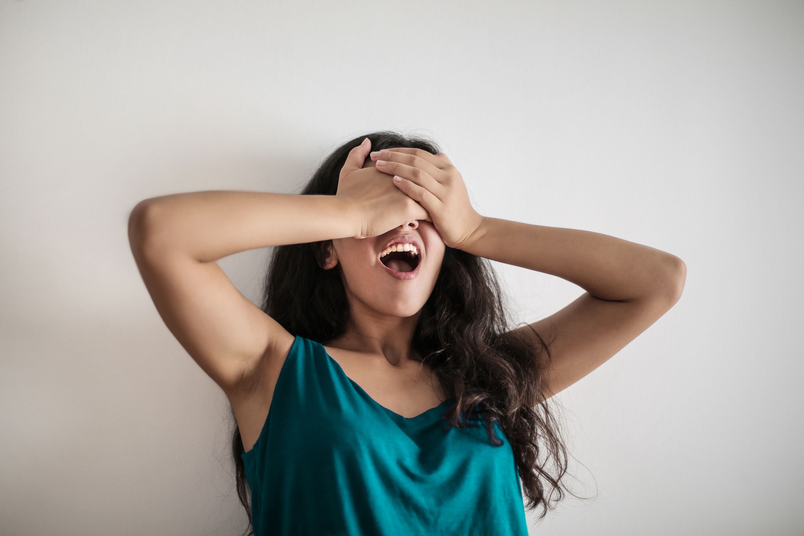 Young long haired female standing with opened mouth and covering eyes with hands in surprise against white background