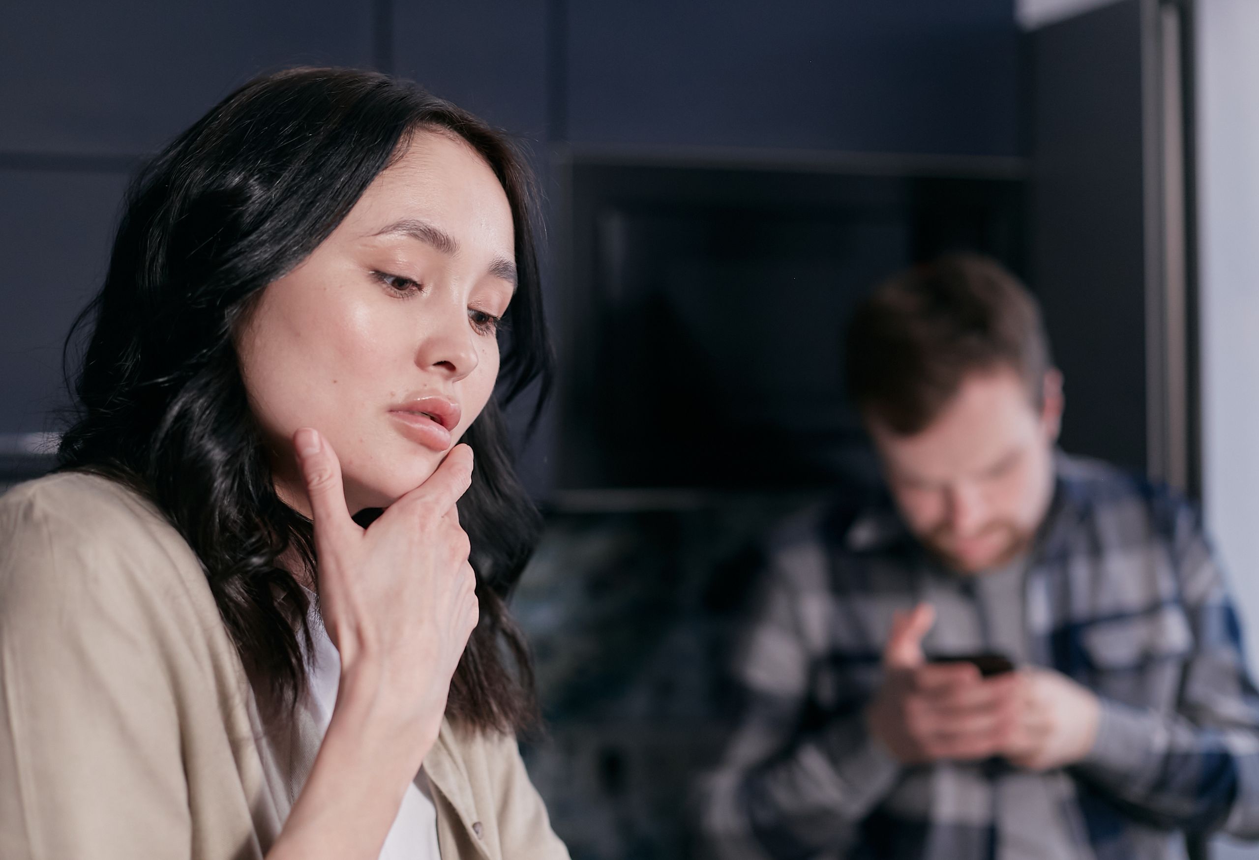 Young woman pondering indoors while man uses smartphone in the background.