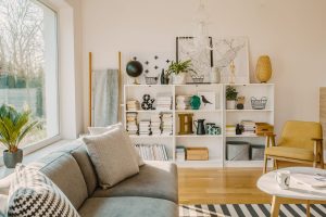 Ladder with blanket standing next to white wooden rack with decorations, books and plants in bright living room interior with grey couch and mustard armchair