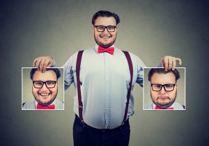 Young cheerful man in glasses holding pictures with good and bad emotions having mood swings and smiling at camera