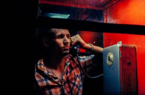 A man of criminal appearance in a payphone booth at night. Talking on the phone with a revolver and a cigarette.