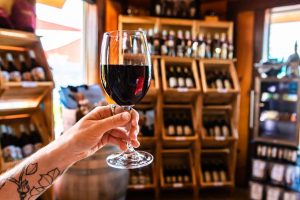 hand with tattoo holding a glass of red wine selective focus close up view against wine bottles on tasting room display wooden racks background