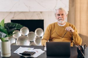 Mature gray-haired man sitting at his workplace at home and looking directly at the camera