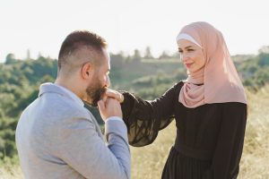 Muslim love story close-up. Mixed couple smiles and hugs on the green hills. Woman weared in hijab looks to her man. Advert for on-line dating agency