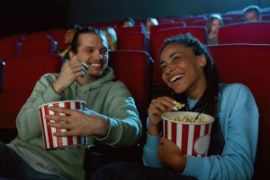 Cheerful young couple laughing, having popcorn while watching movie together, sitting in cinema auditorium