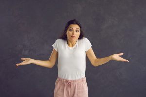 Studio portrait on grey background woman with indifferent face shrugs shoulders to deny or refuse responsibility, pretend dumb or stupid, ignorant of whats happening, clueless about what to do or say