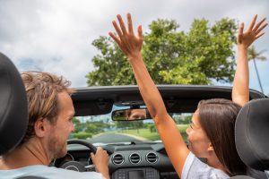 Happy people driving new convertible car on road trip. Asian woman with arms up laughing with boyfriend through open roof having fun driving on summer vacation travel