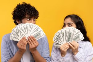 Big win concept. Young indian couple holding and covering faces with a lot of dollar cash fan, yellow studio background