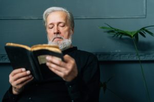 Close-up front view of senior gray-haired aged male reading paper book sitting on chair in dark living room, relax and rest with literature. Handsome mature adult man reading book at nursing home.
