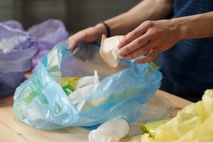 Hand of adolescent guy putting white small plastic yoghurt container into cellophane sack with similar waste while sorting trash in garage