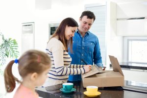 Young happy smiling family three persons unwrapping cardboard cardboard boxes with stuff in light kitchen living room, moving to new flat