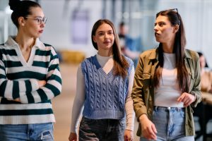 Group of three professional women walking through a modern office hallway, engaged in a casual business conversation, representing teamwork, confidence, and collaboration in the workplace.