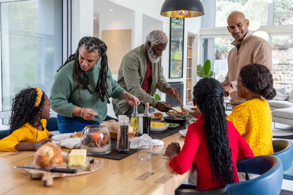 Diverse multigenerational family serving food and passing plates at wooden dining table in kitchen. Openplan, sunlit, contemporary, gathering, sharing, interiors, lifestyle
