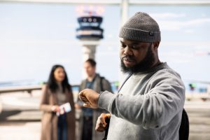 Male traveler checks time on smartwatch in airport terminal, with other passengers and control tower in blurry background. Man looking at wrist watch, being late for flight