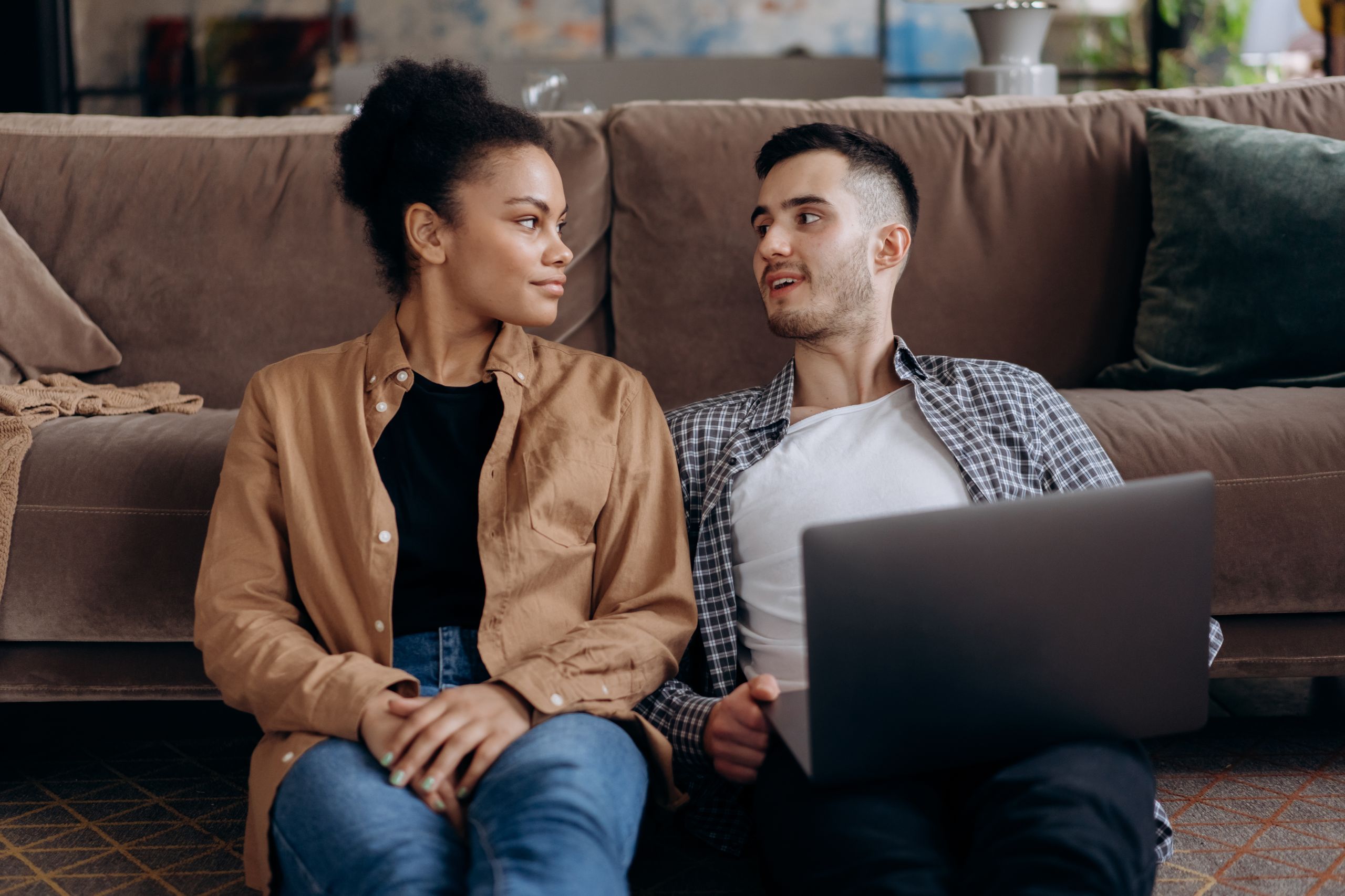 A couple sitting on the floor at home, using a laptop and sharing a moment.
