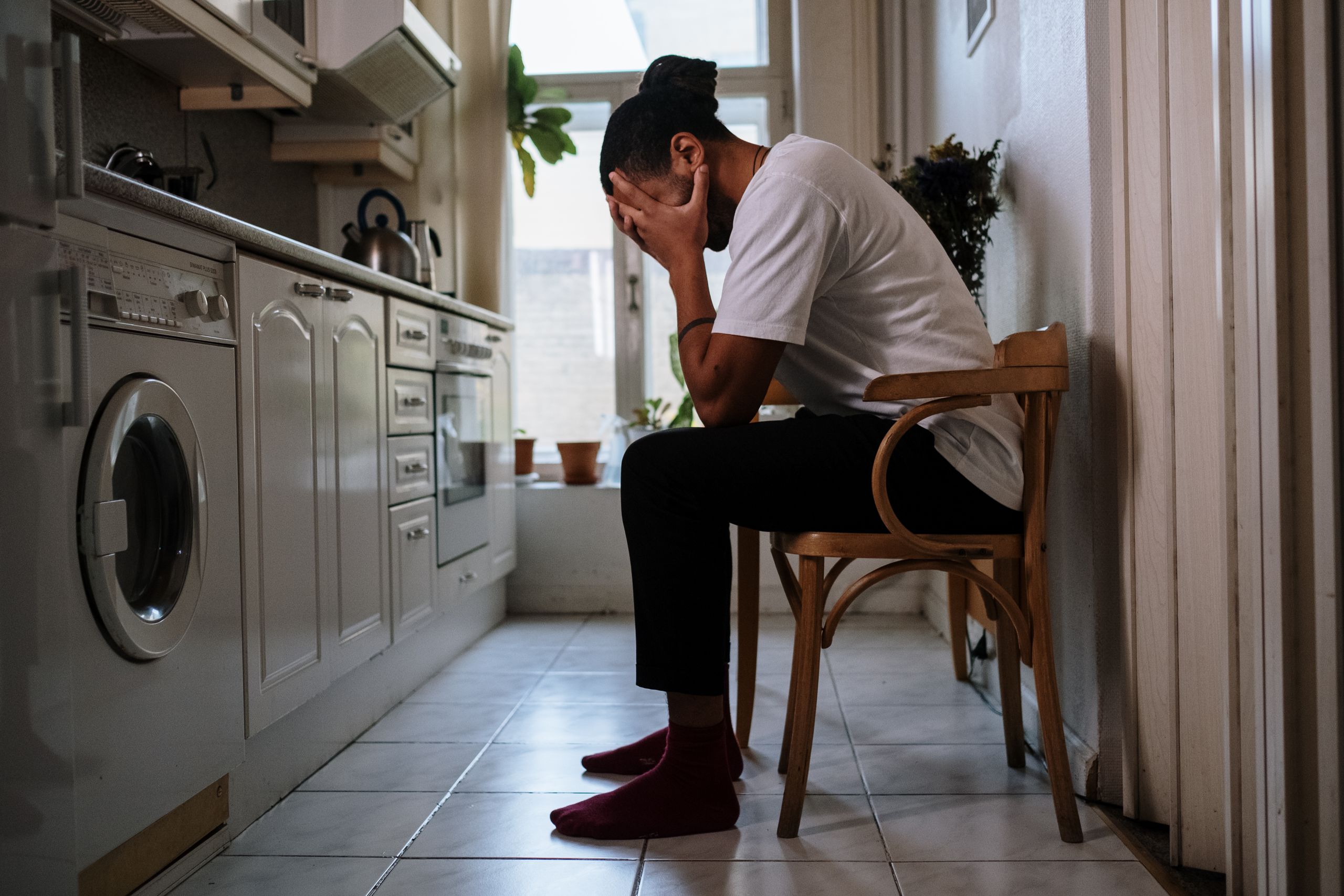 A man sits in a kitchen, visibly stressed with head in hands, capturing an emotional moment indoors.