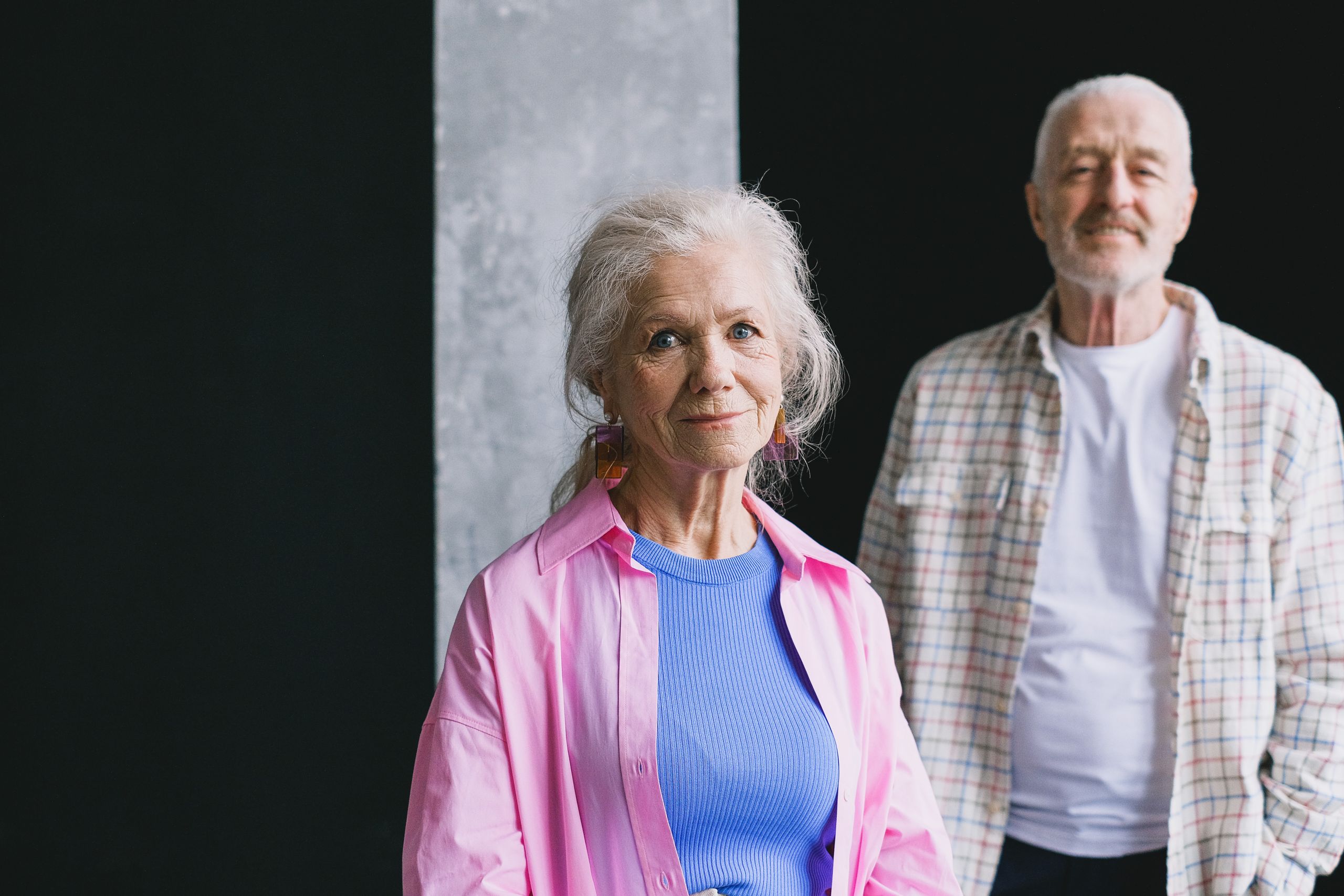 A senior couple smiling warmly indoors, with checkered and pink shirts.