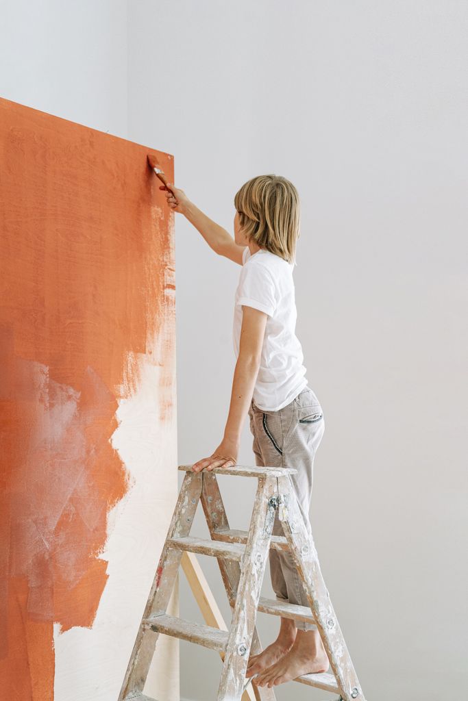 A young boy stands on a ladder painting a wall orange in a bright indoor setting.