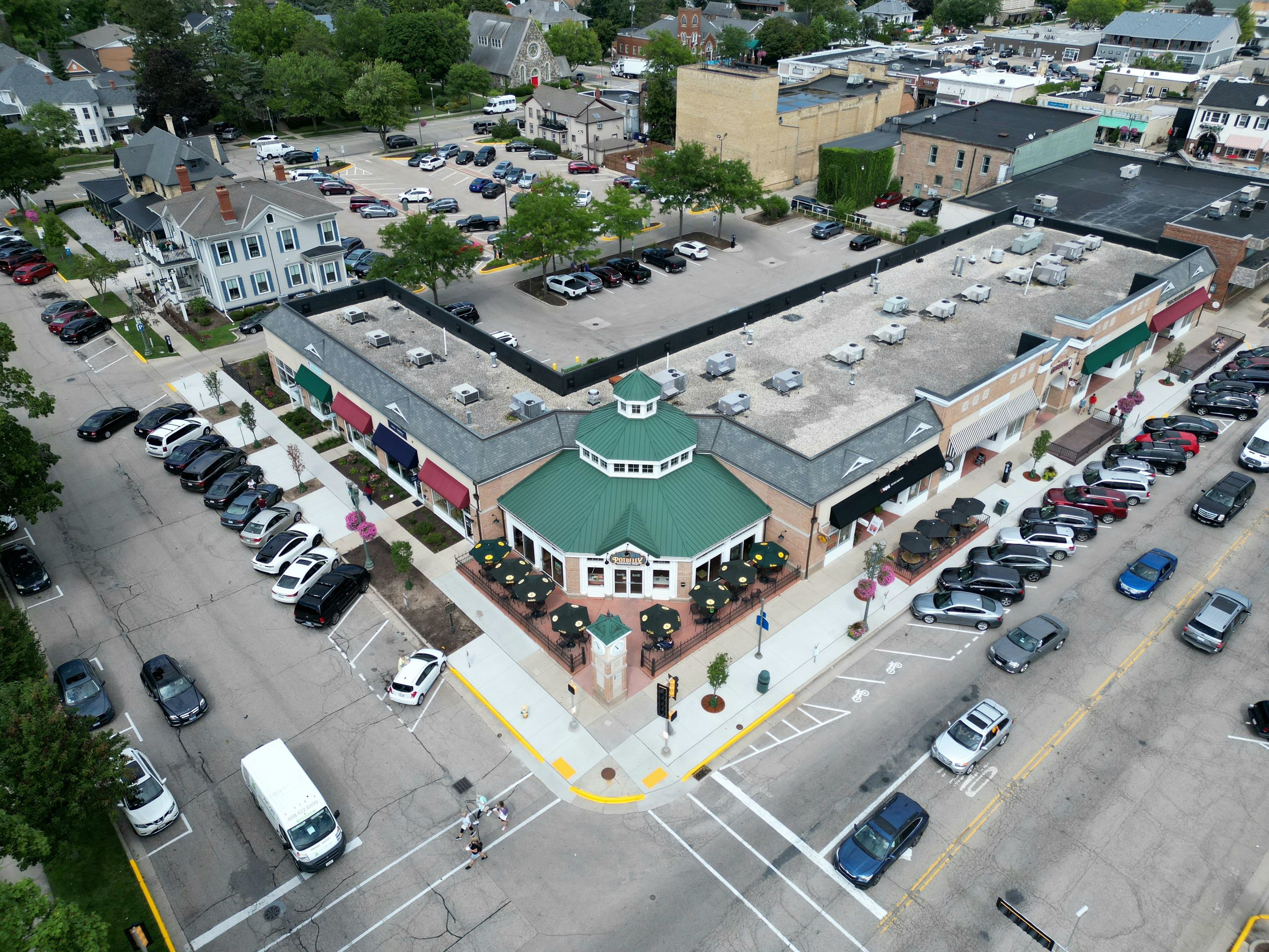 Bird's-eye view of a bustling suburban shopping center with parked cars and tree-lined streets.
