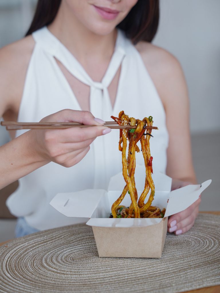 Close-up of a woman eating noodles with chopsticks from a takeout box, showcasing Asian cuisine.