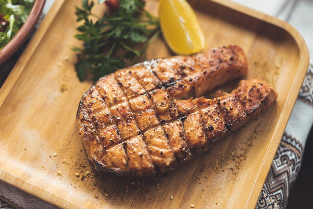 Close-up of seasoned grilled salmon steak with lemon and herbs on a wooden platter.